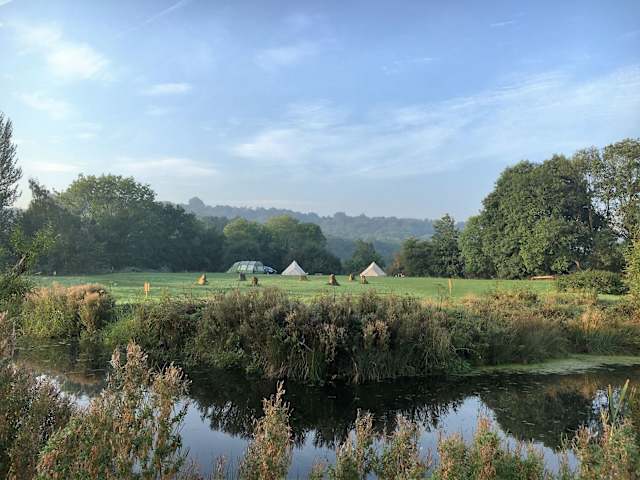 Misty morning view across a pond to a grassy meadow with bell tents and wooded hills beyond