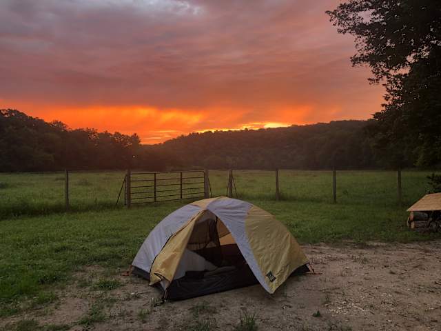 Yellow tent glowing at dusk beneath fiery orange sunset over forested hills and pasture