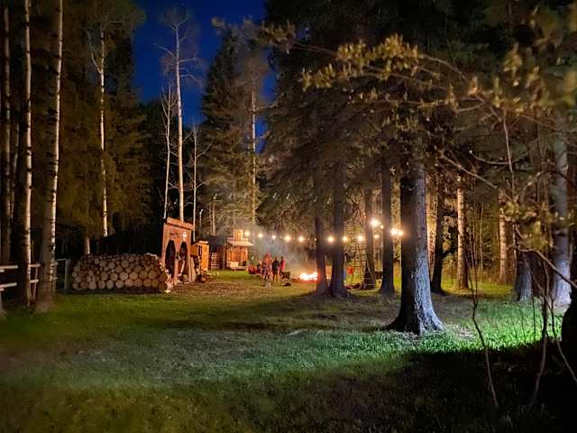 Twilight campsite with string lights, bonfire, log pile and rustic cabin among birch trees