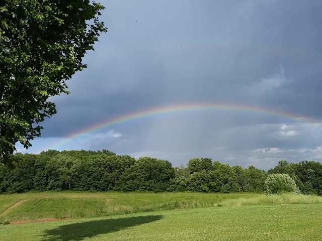 Full rainbow arcing over green fields and trees beneath stormy sky