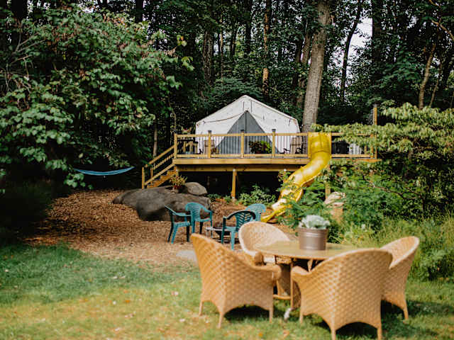 Canvas tent glowing with string lights at dusk above a slide, fire pit with chairs in foreground