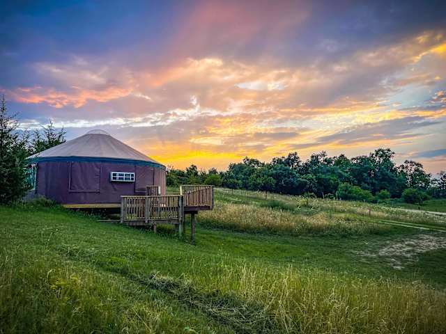 Purple yurt on wooden deck under vivid orange and pink sunset sky, surrounded by grassy field