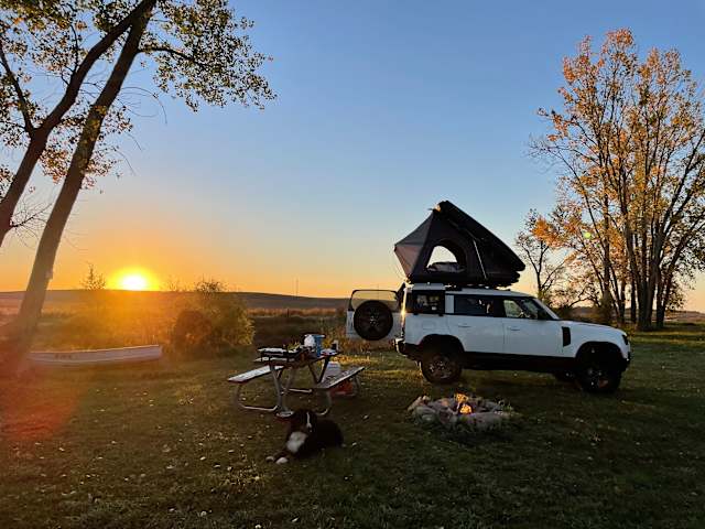 White Land Rover with rooftop tent, dog resting, sunset over prairie fields