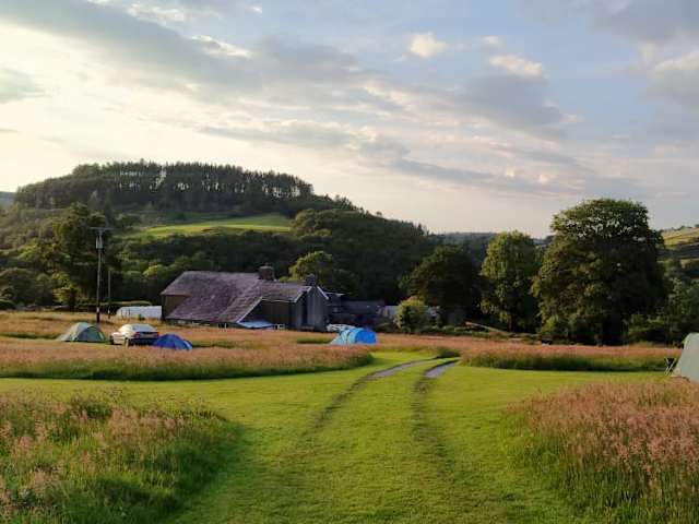 Meadow at golden hour with small tents, stone farmhouse, and wooded hills beyond