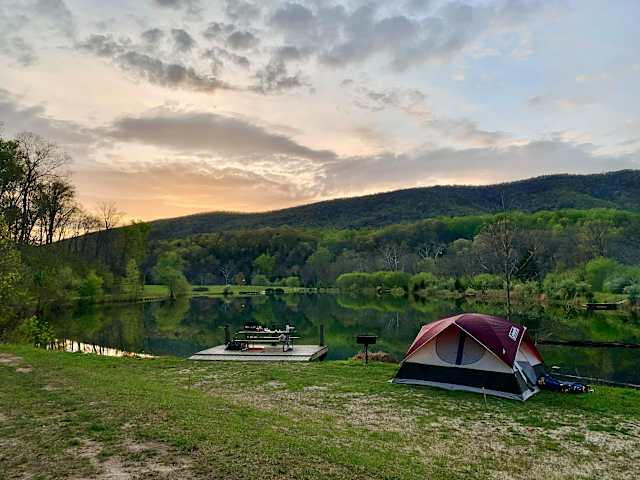 Red tent beside calm lake at sunset with forested mountain reflection and picnic table on dock