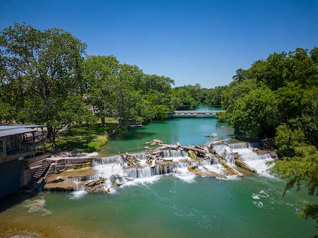 Aerial view of cascading dam waterfall on river lined with lush green trees under blue sky