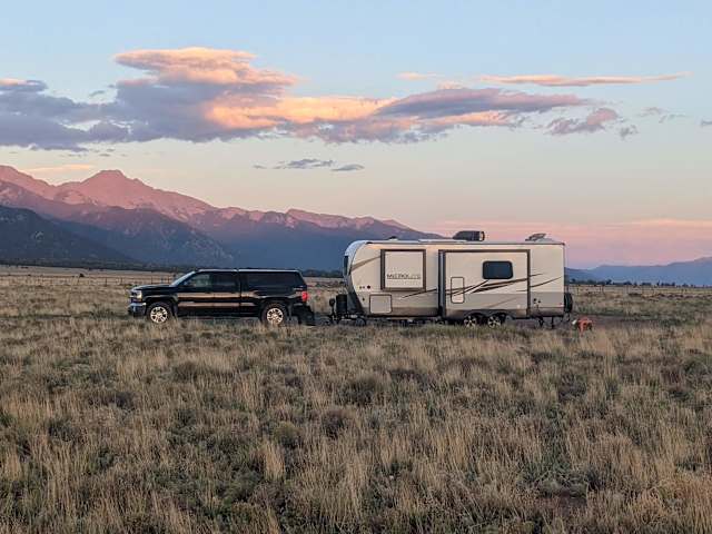 Truck towing travel trailer parked on high desert plain under pink sunset-lit mountain range