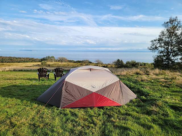 Dome tent pitched on grassy bluff overlooking calm sea under blue sky