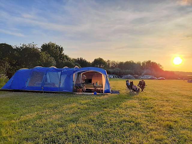 Large blue tent glowing at sunset with campers relaxing in chairs nearby