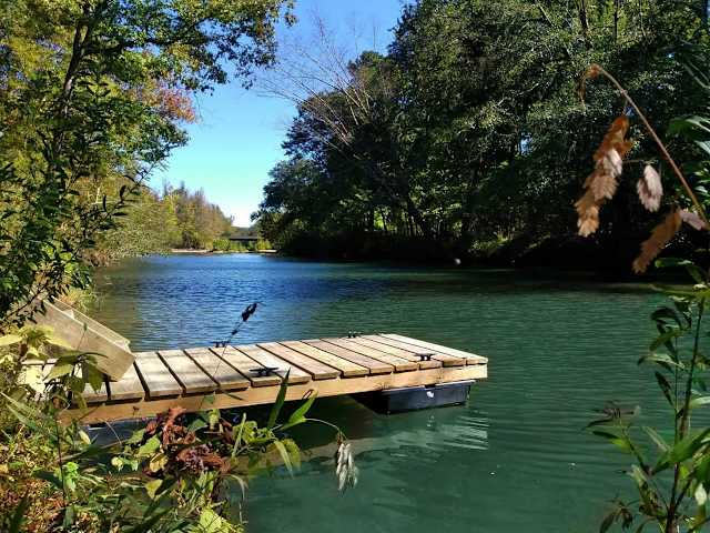 Wooden floating dock extending into calm green river surrounded by dense forest under blue sky