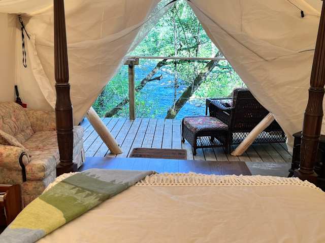 View through open canvas tent flaps toward a river and forest deck with wicker chairs