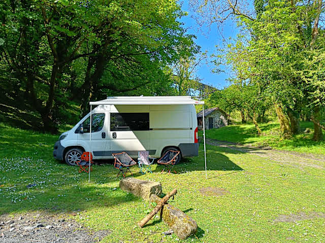 White campervan with awning and folding chairs parked on sunny green meadow among trees