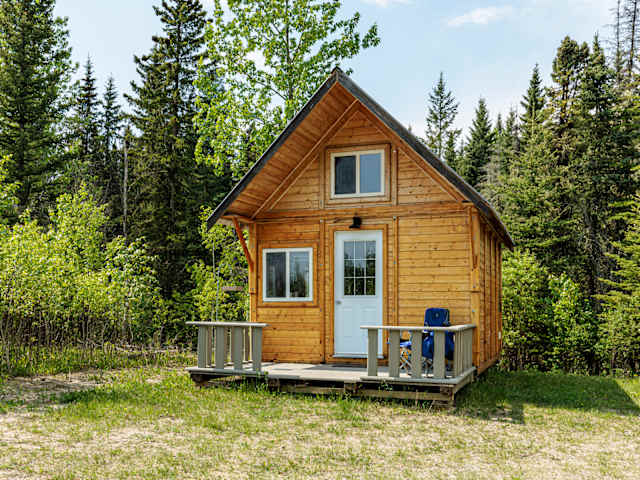 Wooden A-frame cabin with white door and deck surrounded by evergreen forest under blue sky