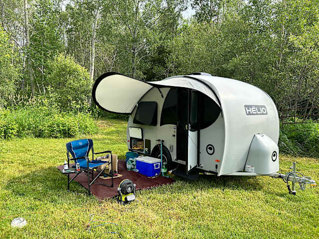 Teardrop trailer named Helio with awning open, camp chairs and gear on mat, surrounded by trees