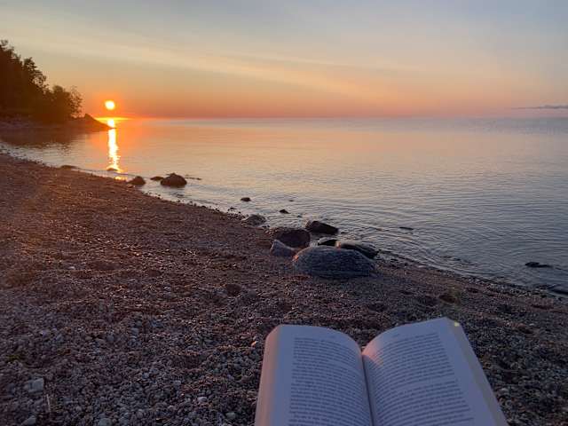 Open book on pebble beach facing a glowing sunrise over calm lake water