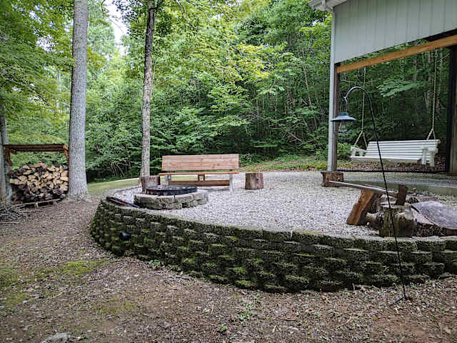 Stone-walled fire pit patio with wooden bench and swing under trees near pole barn
