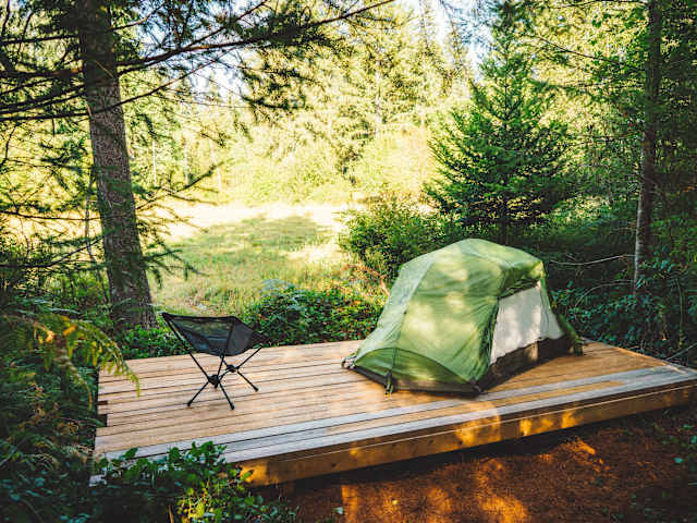 Person sits in camp chair on wooden platform beside green tent overlooking sunlit meadow