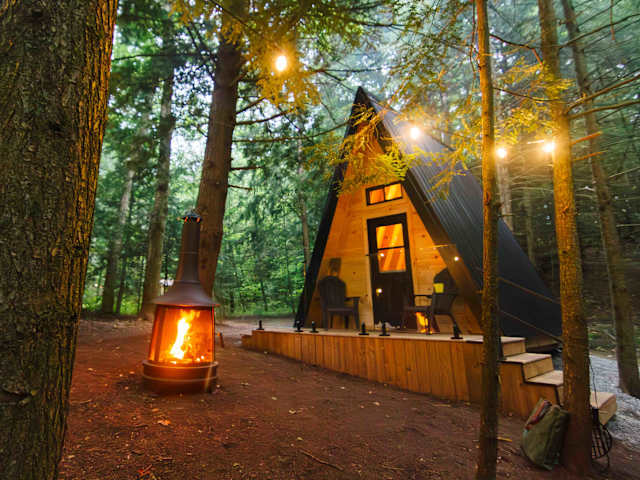 A-frame cabin glowing at dusk in forest with chiminea fire and string lights