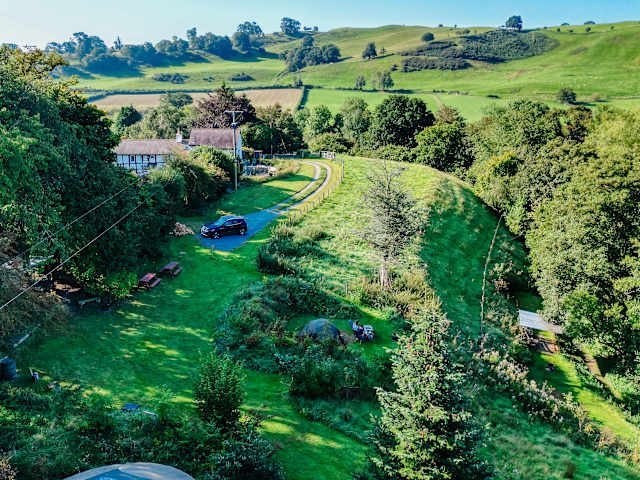 Aerial view of green valley campsite with cottage, winding path, tents and rolling hills