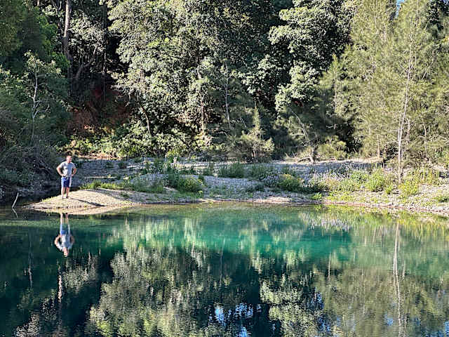 Person standing at edge of turquoise lagoon surrounded by forest, reflection in still water