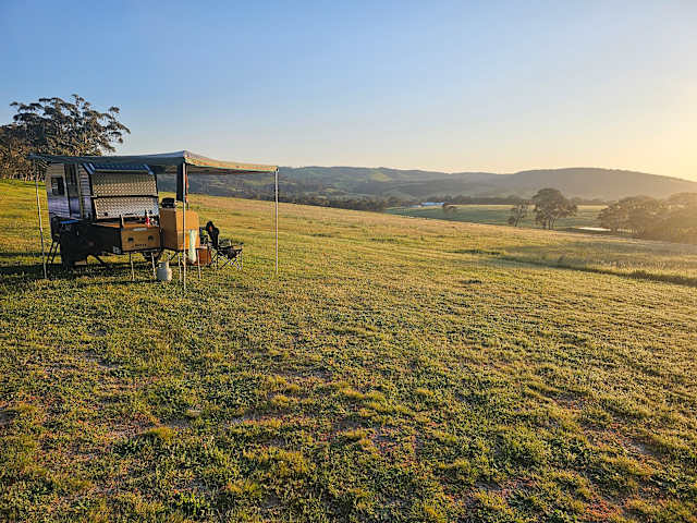 Camper trailer with awning set up on grassy hillside at sunset, rolling hills in distance