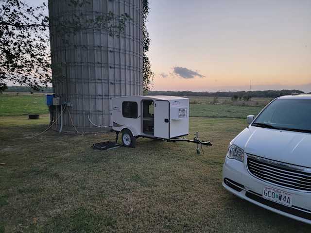 Small teardrop trailer parked beside a tall grain silo at sunset with open field beyond