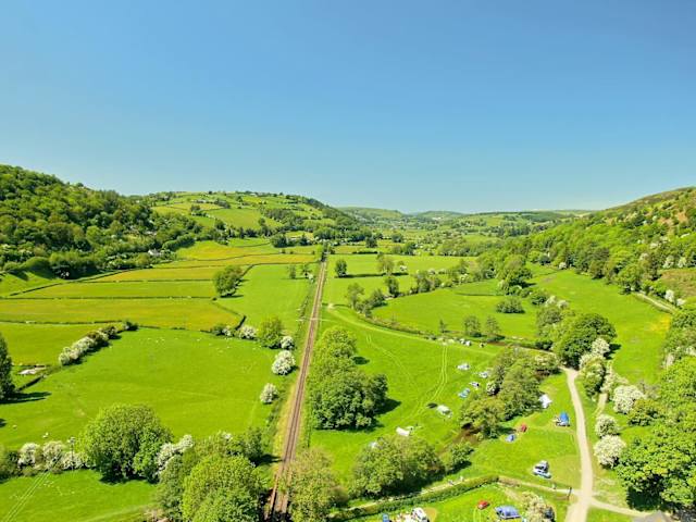 Aerial view of green valley with railway line, fields, river, and campsite tucked among trees