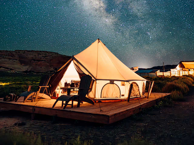 Illuminated glamping tent on wooden deck under starry sky with visible Milky Way, desert cliffs behind