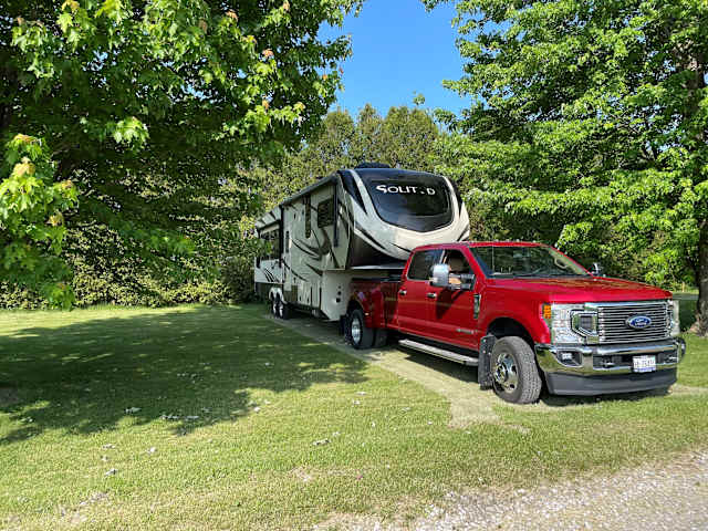 Red pickup truck towing a large fifth-wheel trailer parked on green grass under trees, clear blue sky