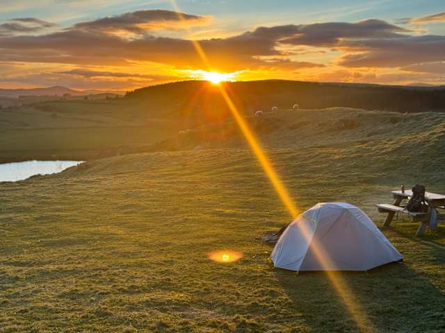 Tent pitched on hillside campsite at sunset with sunburst and rolling hills beyond