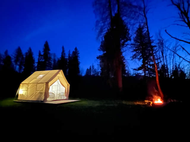 Canvas glamping tent lit warmly at dusk beside a campfire under deep blue twilight sky