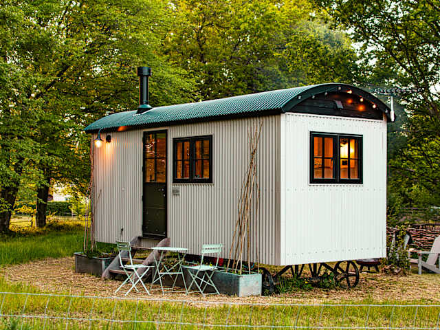 Shepherd's hut with warm interior lights glowing at dusk, surrounded by trees and grass