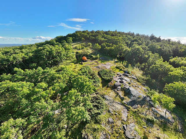 Aerial view of tree-covered mountain ridge with an orange tent nestled at the summit