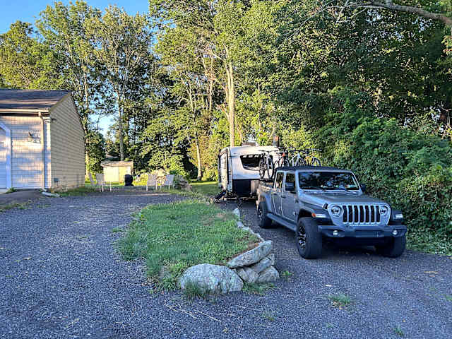 Silver Jeep and travel trailer parked on gravel driveway at dusk with trees behind