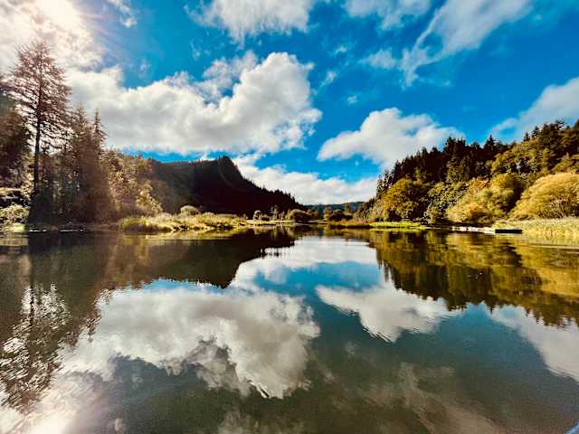 Calm lake reflecting clouds and forested hills under a bright blue sky with sun flare