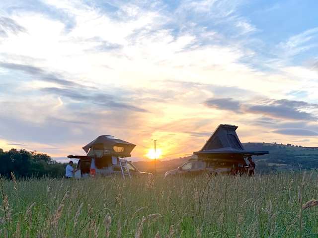 Two camper vehicles with rooftop tents in a field at sunset, golden sky, silhouetted figure