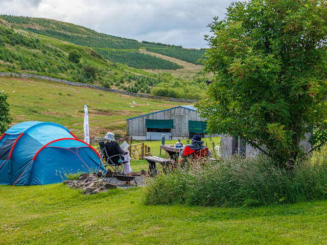 Blue tent with campers seated beside it, green hills and barn in background