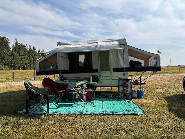 Pop-up trailer with awning extended and camp chairs set up in a grassy field near forest