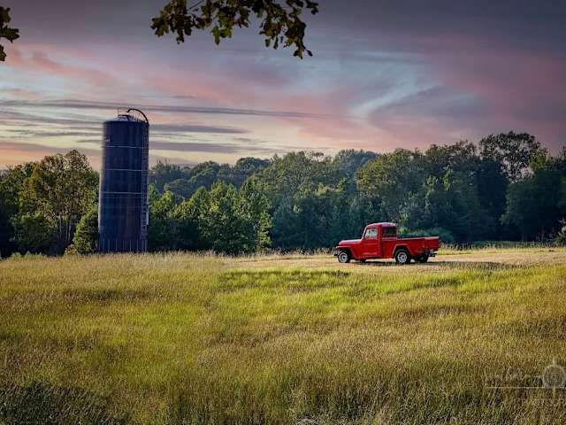 Red vintage pickup truck parked near a tall silo in a grassy field at dusk with colorful sky