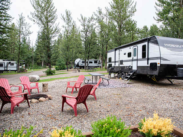 White travel trailer parked at gravel site with red chairs around fire pit among pines