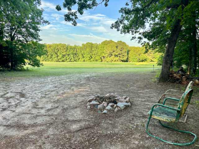 Stone fire pit and vintage chair on dirt clearing framed by trees with green field beyond