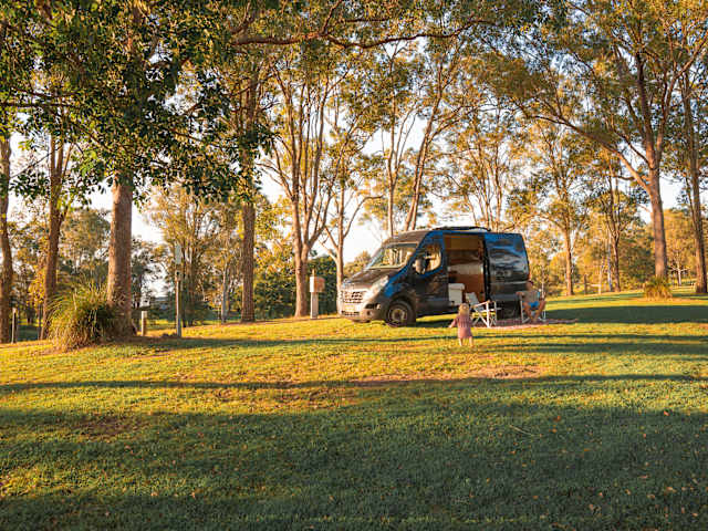 Camper van parked on grass among trees at golden hour, child walking nearby