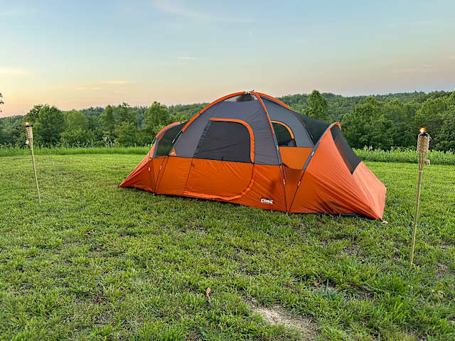 Orange dome tent glowing in grassy field at dusk with torches and tree line