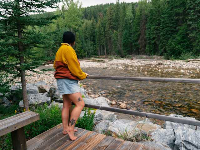 Person standing barefoot on a wooden deck railing overlooking a rocky forest creek