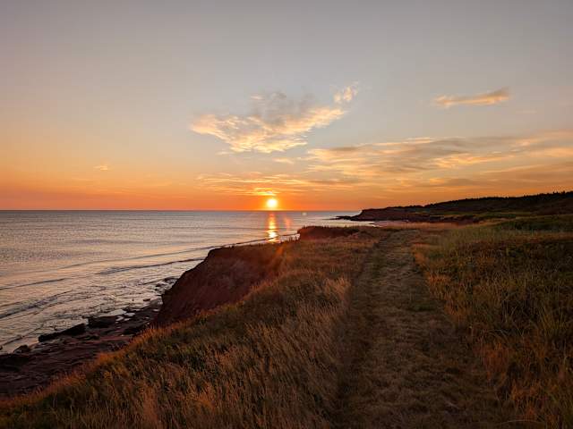 Sun setting over ocean horizon beside red sandstone cliffs and grassy path