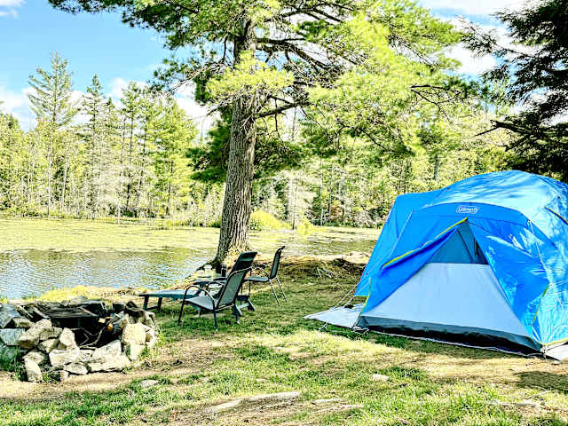 Blue tent and camp chairs beside a fire pit overlooking a still pond