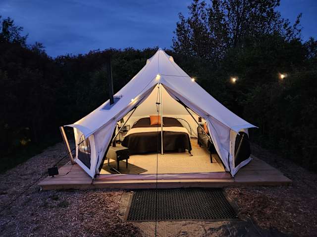 White canvas bell tent glowing warmly at dusk with visible bed and string lights against dark forest
