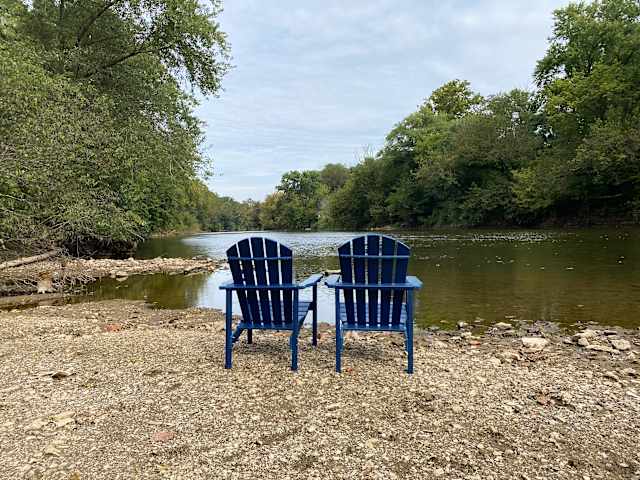 Two blue Adirondack chairs facing a calm tree-lined river under soft sky