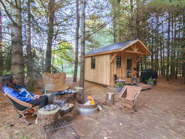 Wooden cabin with yellow door in forest clearing, family gathered around campfire at dusk