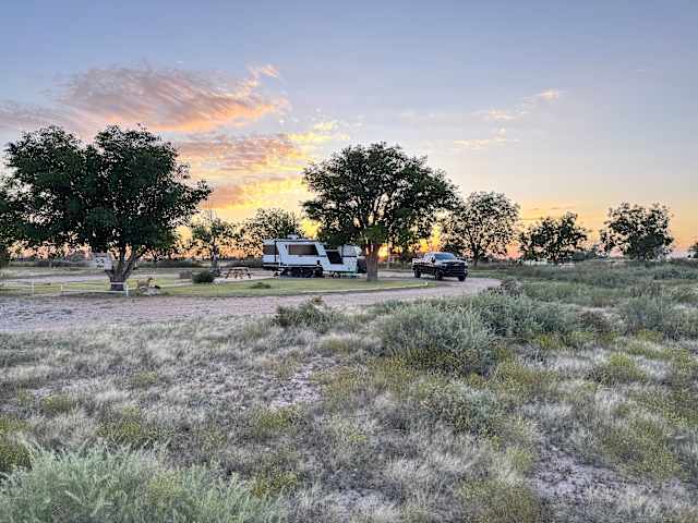 Travel trailer and truck parked among desert trees under a pink and orange sunset sky